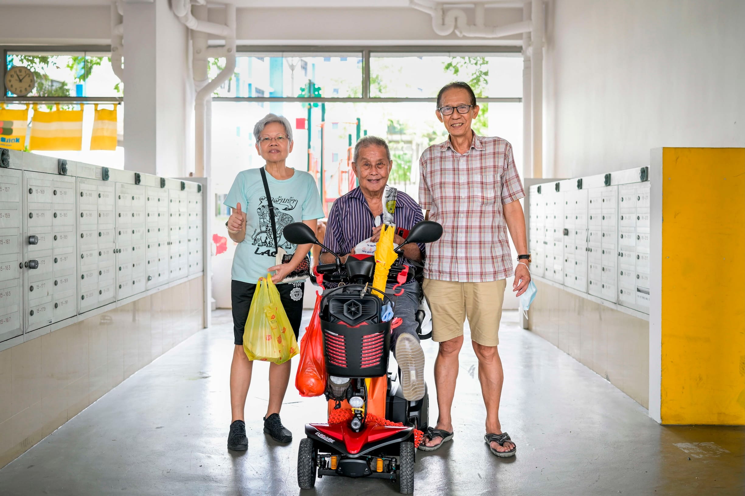 Three older adults pose near mailboxes. One sits on a red EuroCare scooter.